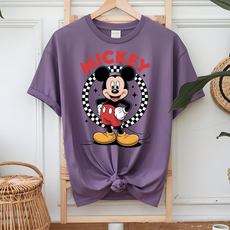 May include: A purple short-sleeve t-shirt featuring a graphic of Mickey Mouse standing inside a black and white checkered circle. The word MICKEY is printed in large red capital letters above the character. The shirt has a crew neckline.