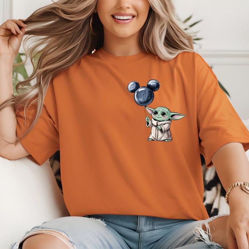 以下が含まれることがあります： 左胸にグラフィックプリントが施されたオレンジ色の半袖Tシャツ。グラフィックには、明るい色のローブを着て、ネズミの耳の形をした濃い青色の風船を持っている、大きな耳をした小さな緑色の生き物が描かれています。
