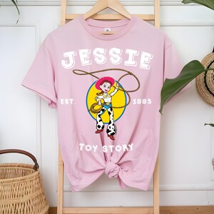 Puede incluir: Camiseta rosa claro con un dise&ntilde;o de Jessie de Toy Story. El dise&ntilde;o incluye el nombre "JESSIE" en letras blancas, el a&ntilde;o "1995" y las palabras "Toy Story". El dise&ntilde;o muestra a Jessie con un lazo.