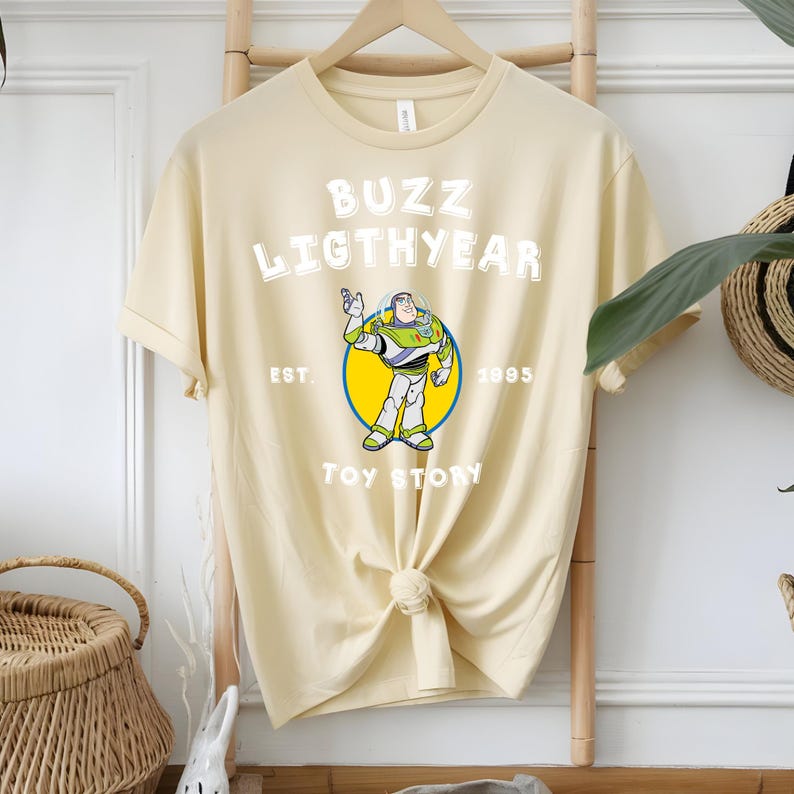 Puede incluir: Camiseta color crema con un dise&ntilde;o de Buzz Lightyear de Toy Story. El dise&ntilde;o incluye el texto "BUZZ LIGHTYEAR" sobre una imagen del personaje, con "EST. 1995" y "TOY STORY" debajo. La camiseta est&aacute; anudada en la parte inferior.