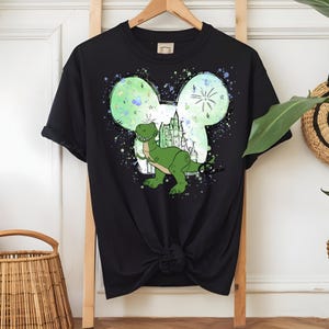 May include: Black t-shirt with a green dinosaur design within a Mickey Mouse ear outline. The graphic includes a castle and fireworks, with a splatter effect. The shirt is displayed on a wooden hanger.