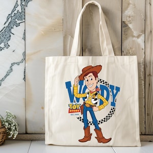 May include: Cream-colored tote bag featuring a cartoon illustration of Woody from Toy Story. The design includes Woody in a cowboy hat, a blue "WOODY" text, and the "TOY STORY EST. 1999" logo. The bag has white handles.