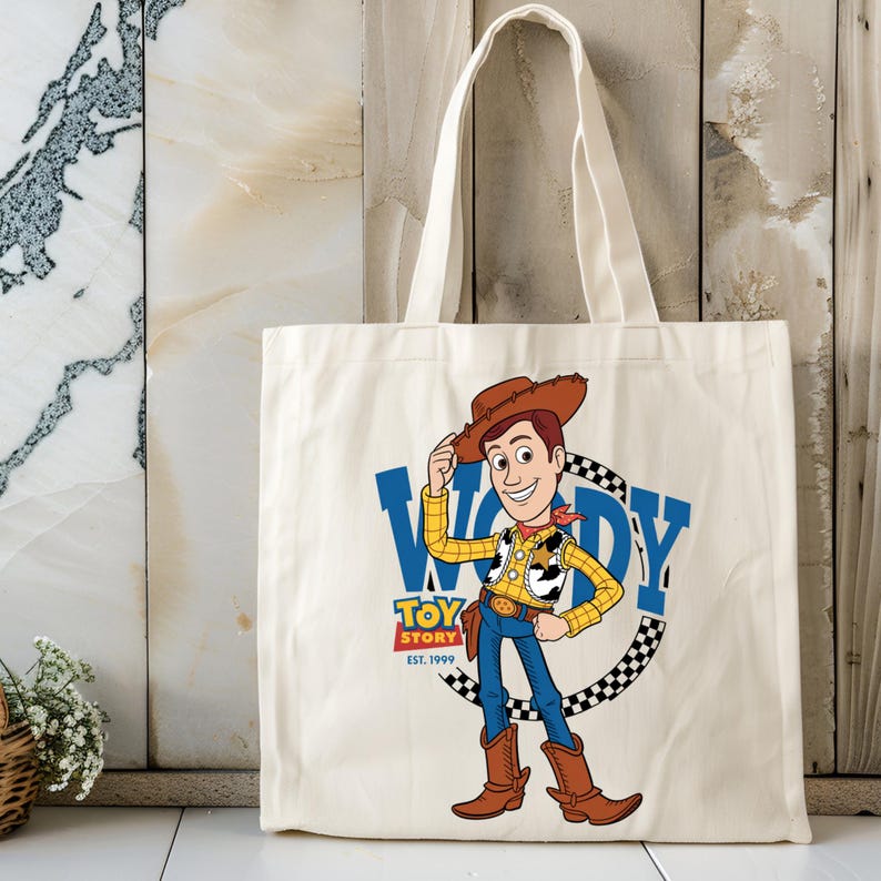 May include: Cream-colored tote bag featuring a cartoon illustration of Woody from Toy Story. The design includes Woody in a cowboy hat, a blue "WOODY" text, and the "TOY STORY EST. 1999" logo. The bag has white handles.