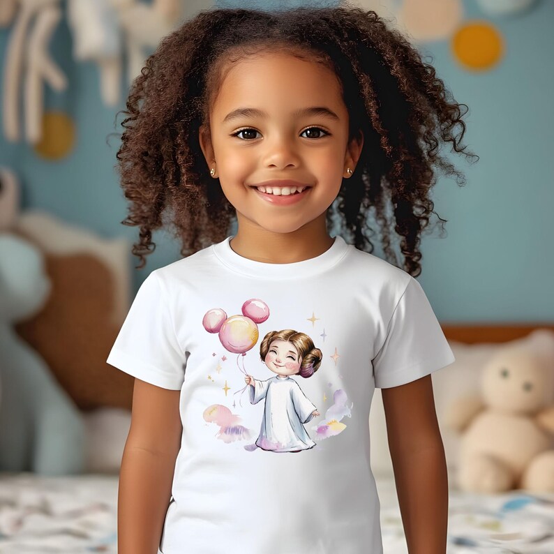 以下が含まれることがあります： 白い半袖Tシャツ。茶色の髪をお団子に結い白いローブを着たキャラクターのグラフィックイラストが描かれており、キャラクターはネズミの耳の形をしたピンクの風船を持ち、小さな金色の星と柔らかな水彩画風の雲に囲まれています。