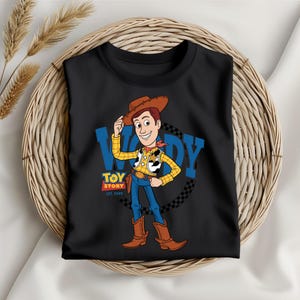 May include: Black t-shirt featuring a cartoon illustration of Woody from Toy Story. Woody is wearing a cowboy hat, vest, and boots. The shirt has the text "WOODY" and "TOY STORY EST. 1995".