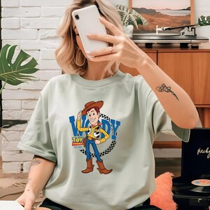 May include: A light green t-shirt featuring a graphic of Woody from Toy Story. The cartoon character is wearing a cowboy hat, yellow shirt, blue jeans, and brown boots. The shirt has the text "WOODY" and "TOY STORY EST. 1995" in a blue and white design.