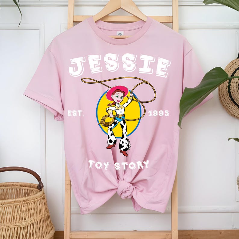 Puede incluir: Camiseta rosa claro con un dise&ntilde;o de Jessie de Toy Story. El dise&ntilde;o incluye el nombre "JESSIE" en letras blancas, el a&ntilde;o "1995" y las palabras "Toy Story". El dise&ntilde;o muestra a Jessie con un lazo.