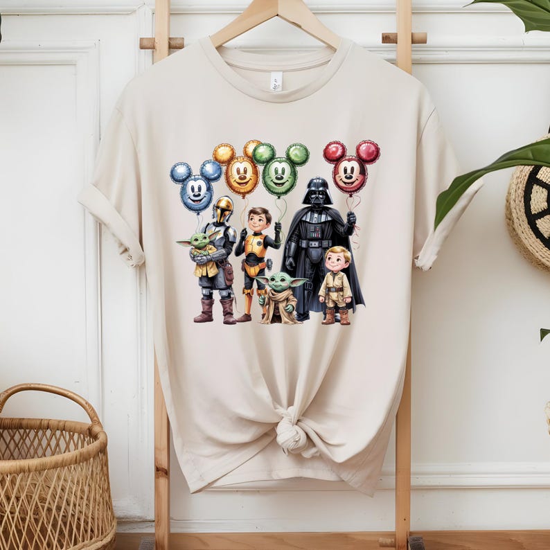 以下が含まれることがあります： ベージュの半袖Tシャツ。マンダロリアン、子供、ダース・ベイダー、小さな男の子などのキャラクターが描かれたグラフィックプリントがあり、全員が青、黄、緑、赤のミッキーマウスの形をしたカラフルな風船を持っています。