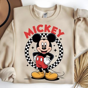 May include: A beige crewneck sweatshirt featuring a graphic of Mickey Mouse standing inside a black and white checkered circle. The word MICKEY is printed in large red letters above the character. The design includes small four-pointed star accents.