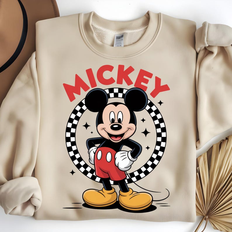 May include: A beige crewneck sweatshirt featuring a graphic of Mickey Mouse standing inside a black and white checkered circle. The word MICKEY is printed in large red letters above the character. The design includes small four-pointed star accents.