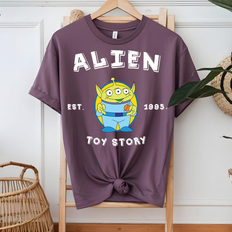 Puede incluir: Camiseta de color malva con un dise&ntilde;o del personaje Alien de Toy Story. El dise&ntilde;o incluye la palabra "ALIEN" encima del personaje, con "EST. 1995" y "TOY STORY" debajo. El Alien es verde, azul y amarillo.