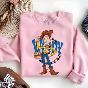May include: A light pink sweatshirt featuring a graphic of Woody from Toy Story. The design includes Woody in his cowboy attire, the words "WOODY" in blue, and "TOY STORY EST. 1999". The sweatshirt has a crew neck and long sleeves.