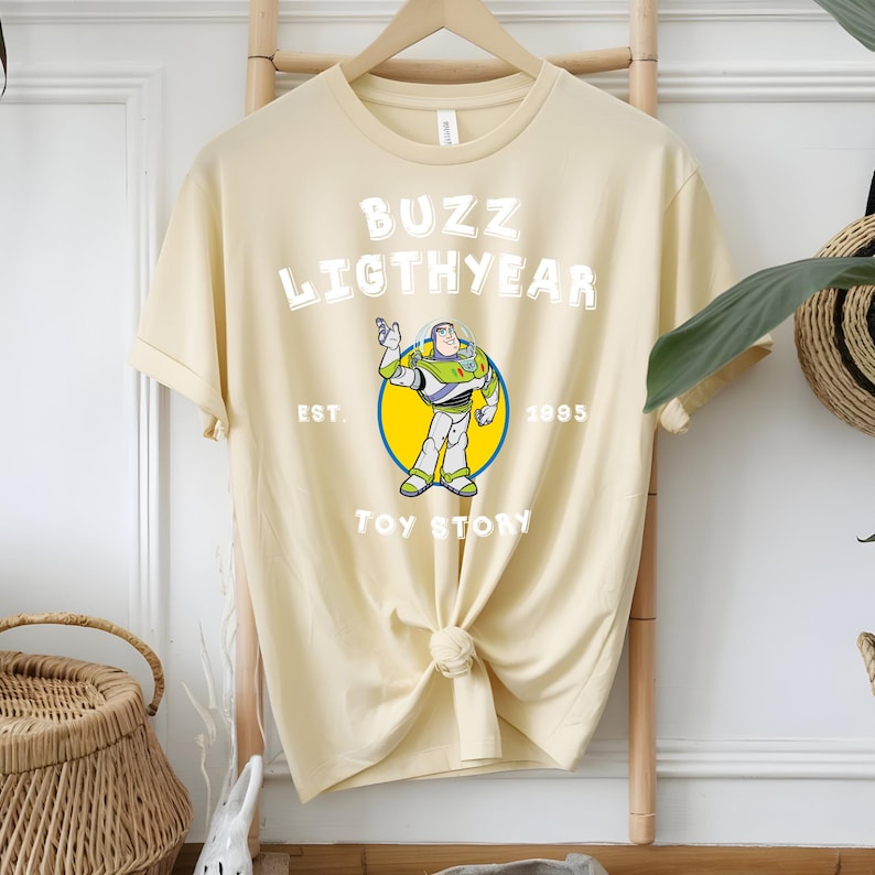 Puede incluir: Camiseta color crema con un dise&ntilde;o de Buzz Lightyear de Toy Story. El dise&ntilde;o incluye el texto "BUZZ LIGHTYEAR" sobre una imagen del personaje, con "EST. 1995" y "TOY STORY" debajo. La camiseta est&aacute; anudada en la parte inferior.