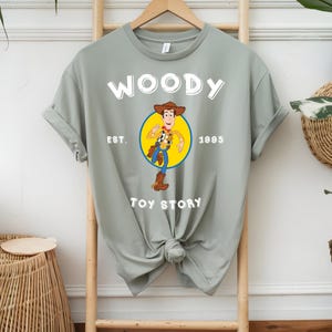 Puede incluir: Una camiseta verde claro con la palabra "WOODY" en blanco sobre una imagen de dibujos animados de Woody de Toy Story, dentro de un c&iacute;rculo amarillo. El texto "EST. 1995" y "TOY STORY" tambi&eacute;n est&aacute;n impresos en la camiseta.
