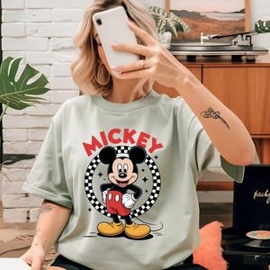 May include: A light sage green t-shirt featuring a graphic print of Mickey Mouse standing inside a black and white checkered circle. The word MICKEY is printed in bold red capital letters above the circular design, which includes small star accents.
