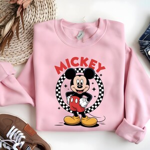 May include: A light pink crewneck sweatshirt featuring a graphic of Mickey Mouse standing in front of a black and white checkered circle. The word MICKEY is printed in red capital letters above the character, accompanied by small black star icons.