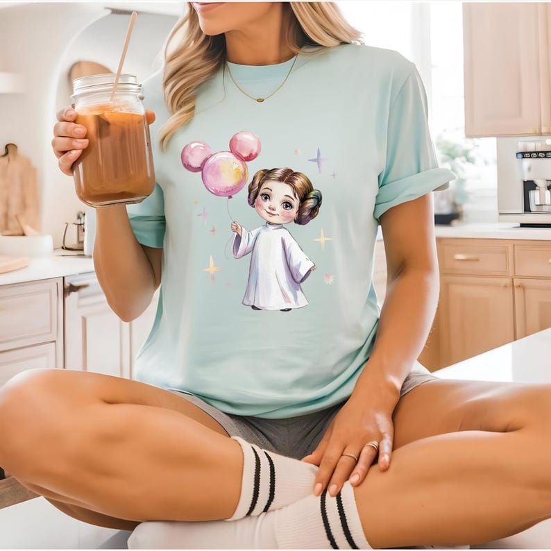 以下が含まれることがあります： 水色のTシャツ。お団子ヘアのキャラクターが白いローブをまとい、ネズミの耳の形をしたピンクの風船を持っているグラフィックイラストが描かれており、周囲には小さなカラフルな星のアクセントが散りばめられています。