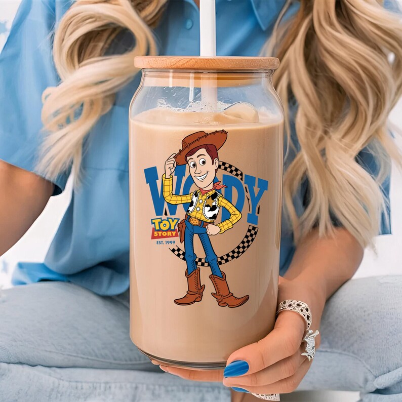 May include: A clear glass tumbler with a wooden lid and a white straw, filled with a light brown beverage. The tumbler features a cartoon image of Woody from Toy Story. The Woody graphic is surrounded by a black and white checkered pattern.