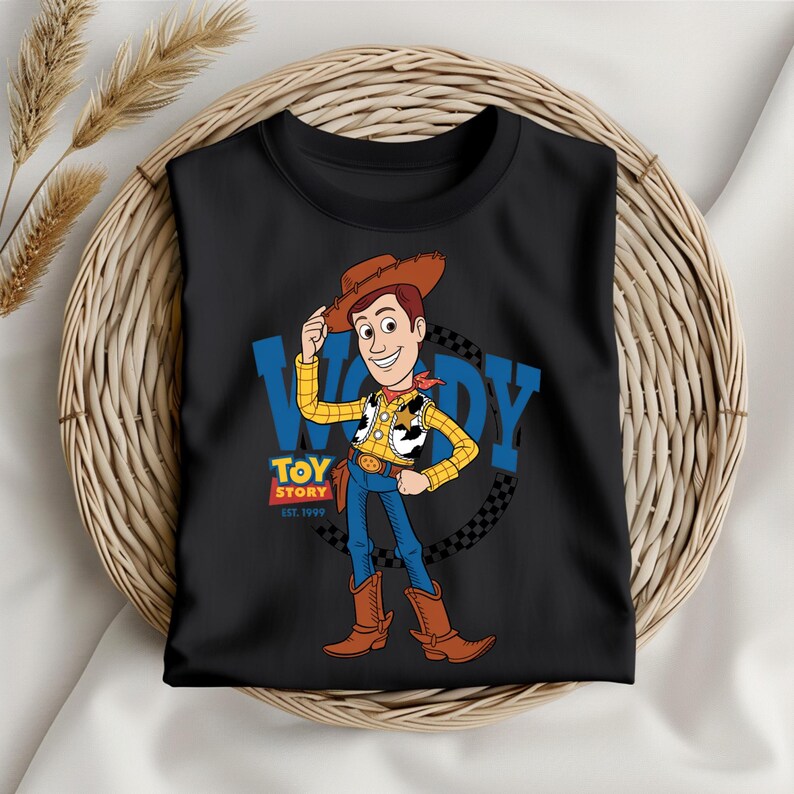 May include: Black t-shirt featuring a cartoon illustration of Woody from Toy Story. Woody is wearing a cowboy hat, yellow shirt, blue jeans, and brown boots. The shirt has the text "WOODY" and "TOY STORY EST. 1995".