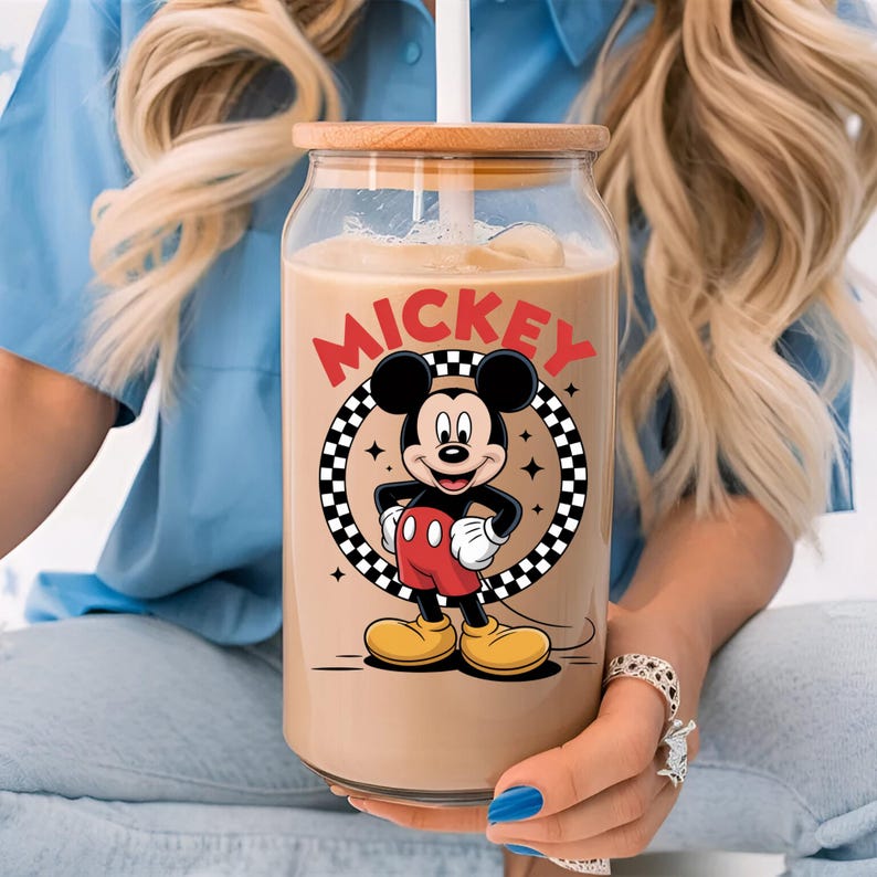 May include: A clear glass tumbler with a bamboo lid and a white straw, filled with a light brown beverage. The glass features a graphic of Mickey Mouse inside a black and white checkered circle, with the red text MICKEY printed above the character.