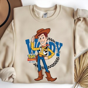 May include: Beige sweatshirt featuring a cartoon illustration of Woody from Toy Story. Woody is wearing a cowboy hat, yellow shirt, blue jeans, and brown boots. The sweatshirt has the words "WOODY" and "TOY STORY EST. 1999" in blue.
