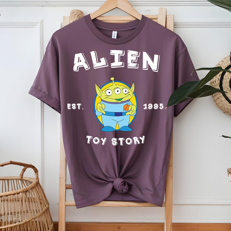 Puede incluir: Camiseta de color malva con un dise&ntilde;o del personaje Alien de Toy Story. El dise&ntilde;o incluye la palabra "ALIEN" encima del personaje, con "EST. 1995" y "TOY STORY" debajo. El Alien es verde, azul y amarillo.