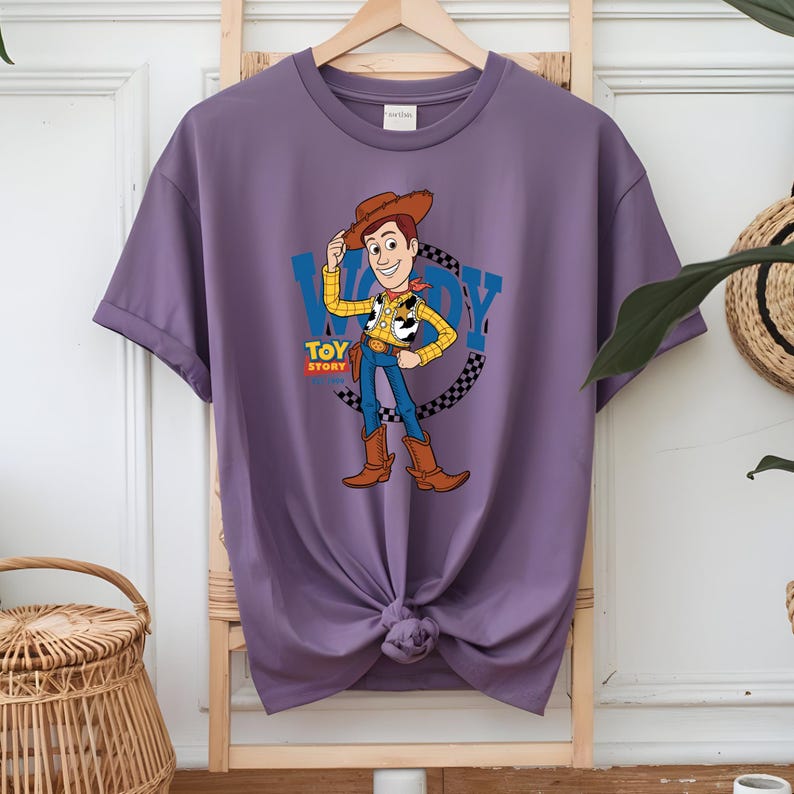 May include: A purple t-shirt featuring a graphic of the character Woody from Toy Story. Woody is wearing a cowboy hat, yellow shirt, blue jeans, and brown boots. The word "WOODY" is in blue letters above the character, with "TOY STORY" below.