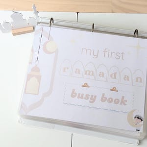 Peut inclure: Un classeur transparent avec des pages affichant le texte "my first ramadan busy book" dans une palette de couleurs douces et neutres. &Eacute;l&eacute;ments d&eacute;coratifs : croissants de lune, &eacute;toiles et silhouette de mosqu&eacute;e.