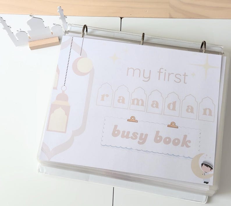 Peut inclure: Un classeur transparent avec des pages affichant le texte "my first ramadan busy book" dans une palette de couleurs douces et neutres. &Eacute;l&eacute;ments d&eacute;coratifs : croissants de lune, &eacute;toiles et silhouette de mosqu&eacute;e.