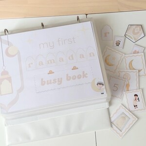 Peut inclure: Un classeur blanc intitul&eacute; "my first Ramadan busy book" avec des accents beiges et dor&eacute;s. Le classeur ouvert r&eacute;v&egrave;le des pages et des cartes d&eacute;tach&eacute;es avec des illustrations de lunes, de lanternes et de personnages. Th&egrave;me : activit&eacute; &eacute;ducative pour enfants.