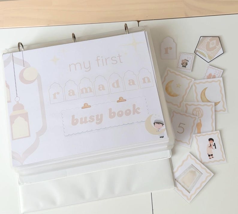Peut inclure: Un classeur blanc intitul&eacute; "my first Ramadan busy book" avec des accents beiges et dor&eacute;s. Le classeur ouvert r&eacute;v&egrave;le des pages et des cartes d&eacute;tach&eacute;es avec des illustrations de lunes, de lanternes et de personnages. Th&egrave;me : activit&eacute; &eacute;ducative pour enfants.