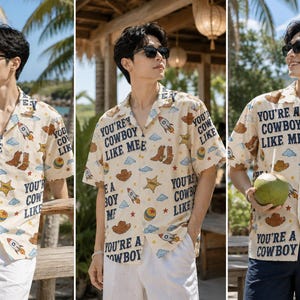 May include: A short-sleeved button-down shirt in a light cream color featuring a repeating pattern of brown cowboy hats, boots, rockets, stars, and blue clouds. The text "YOU'RE A COWBOY LIKE ME" is printed in dark blue across the fabric.
