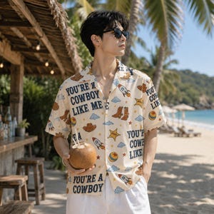 May include: A short-sleeved button-down shirt in cream featuring a repeating pattern of rockets, cowboy boots, sheriff stars, clouds, and balls, with the text YOU'RE A COWBOY LIKE ME printed in dark blue lettering across the fabric.
