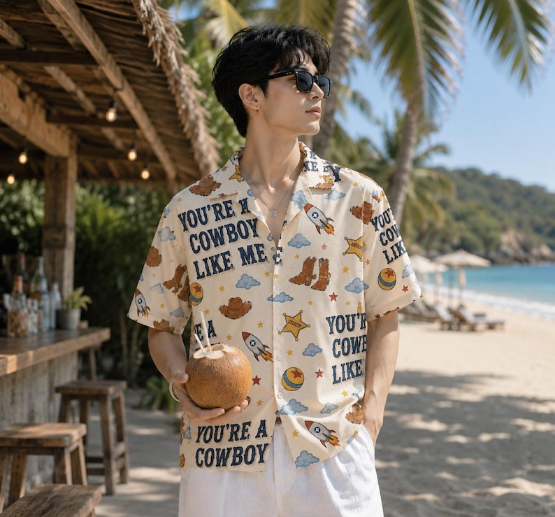 May include: A short-sleeved button-down shirt in cream featuring a repeating pattern of rockets, cowboy boots, sheriff stars, clouds, and balls, with the text YOU'RE A COWBOY LIKE ME printed in dark blue lettering across the fabric.