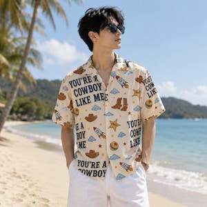 May include: A short-sleeved button-down shirt in a light beige color featuring a repeating pattern of brown cowboy hats, boots, sheriff stars, rockets, blue clouds, red stars, and yellow balls, with the text "YOU'RE A COWBOY LIKE ME" printed in black.