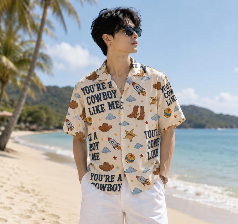 May include: A short-sleeved button-down shirt in a light beige color featuring a repeating pattern of brown cowboy hats, boots, sheriff stars, rockets, blue clouds, red stars, and yellow balls, with the text "YOU'RE A COWBOY LIKE ME" printed in black.