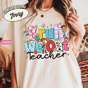 May include: Ivory-colored t-shirt with the words "Bruh We Out Teacher" in colorful, playful lettering. The design includes cartoon characters, flowers, and stars. The shirt is from Comfort Colors.
