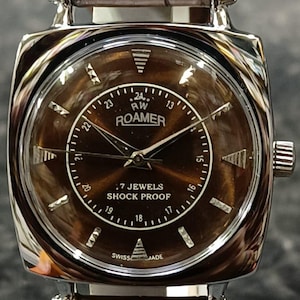 Könnte beinhalten: Eine Vintage-Roamer-Uhr mit braunem Zifferblatt und Edelstahlgehäuse. Die Uhr hat ein braunes Lederarmband und den Text "7 JEWELS SHOCK PROOF" und "SWISS MADE". Die Uhr hat ein quadratisches Zifferblatt und einen Sekundenzeiger.