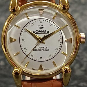Może przedstawiać: Złoty zegarek Roamer z białą tarczą, czarnymi cyframi i wskazówkami. Zegarek ma brązowy skórzany pasek. Na tarczy widnieje napis "RW ROAMER", "17 JEWELS" i "SHOCK PROOF".