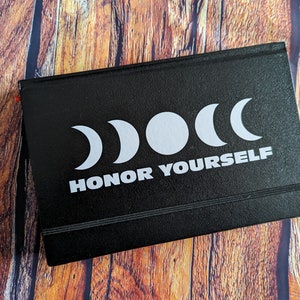 May include: Black journal with a white design of three crescent moons and the text "HONOR YOURSELF".