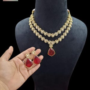 Op de afbeelding: Een goudkleurige ketting met twee lagen, voorzien van driehoekige witte stenen en een centrale druppelvormige rode hanger. De set bevat bijpassende oorbellen, elk met een driehoekige witte steen en een grotere druppelvormige rode steen.