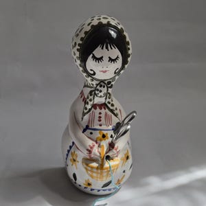 Könnte beinhalten: Keramikfigur einer Frau mit einem Kopftuch mit schwarzen Punkten. Die Figur hat geschlossene Augen und hält einen Korb mit gelben Blumen und einer Schere. Die Schürze der Frau ist mit gelben Blumen verziert.