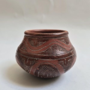 May include: A small, round, brown ceramic pot with carved, wave-like patterns. The pot has a wide opening and a dark brown color. The carved designs are lighter in color, creating a contrast against the darker background.