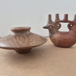 May include: Two terracotta pottery pieces. One is a shallow, wide-brimmed bowl with a small, rounded base. The other is a bird-like sculpture with a long beak, three vertical protrusions, and spiral details. Both pieces are in shades of brown.