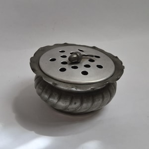May include: A small, antique silver-coloured container with a decorative, ridged body and a perforated lid. The lid has a scalloped edge and a small acorn-shaped handle. The container is likely for holding flowers or potpourri.