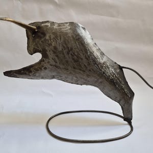 Puede incluir: Escultura metálica de un toro embistiendo. La escultura es principalmente gris con un cuerno dorado. Está montada sobre una base metálica circular, dando la impresión de movimiento y poder. La cola del toro está curvada.