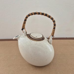 May include: A speckled white ceramic teapot with a brown and tan woven handle. The teapot has a rounded body, a small spout, and a matching lid. The handle is attached with metal hardware. The teapot is a kitchenware item.