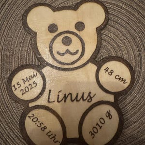 May include: A wooden bear-shaped plaque with the name "Línus" engraved in a cursive font. The plaque includes the date "15 Mai 2025", time "20:58 uhr", weight "3010 g", and height "48 cm". The bear's outline is dark brown.