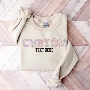 May include: A beige sweatshirt with the word "CUSTOM" in a colorful floral pattern and the words "TEXT HERE" in black block letters. The sweatshirt is folded on a wooden surface.
