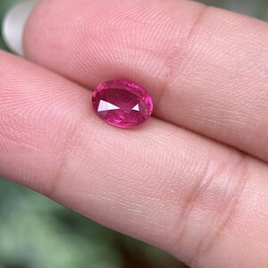 May include: A faceted, oval-cut, ruby-red gemstone. The gem is held between fingers, highlighting its brilliant cut and vibrant colour. The stone's facets reflect light, creating a sparkling effect. The background is blurred, emphasising the gem's details.